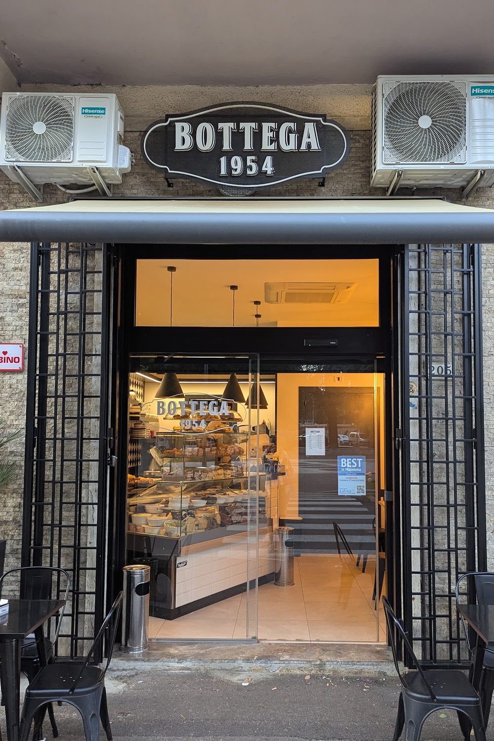 La vetrina di Bottega 1954 in Viale della Pace 205, con l'insegna nera e i tavolini sul marciapiede